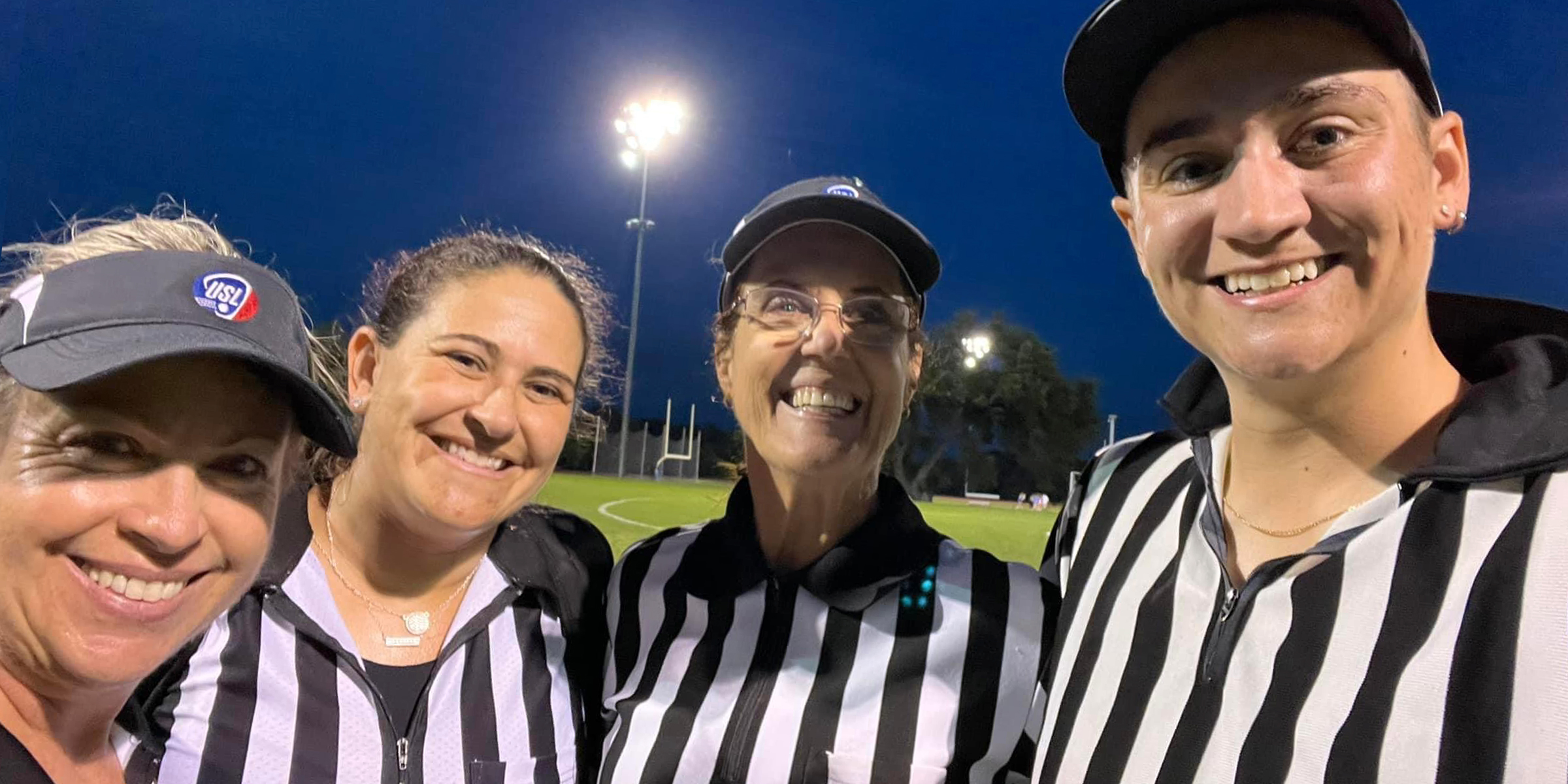photo of four lacrosse officials standing in front of a field and smiling at the camera