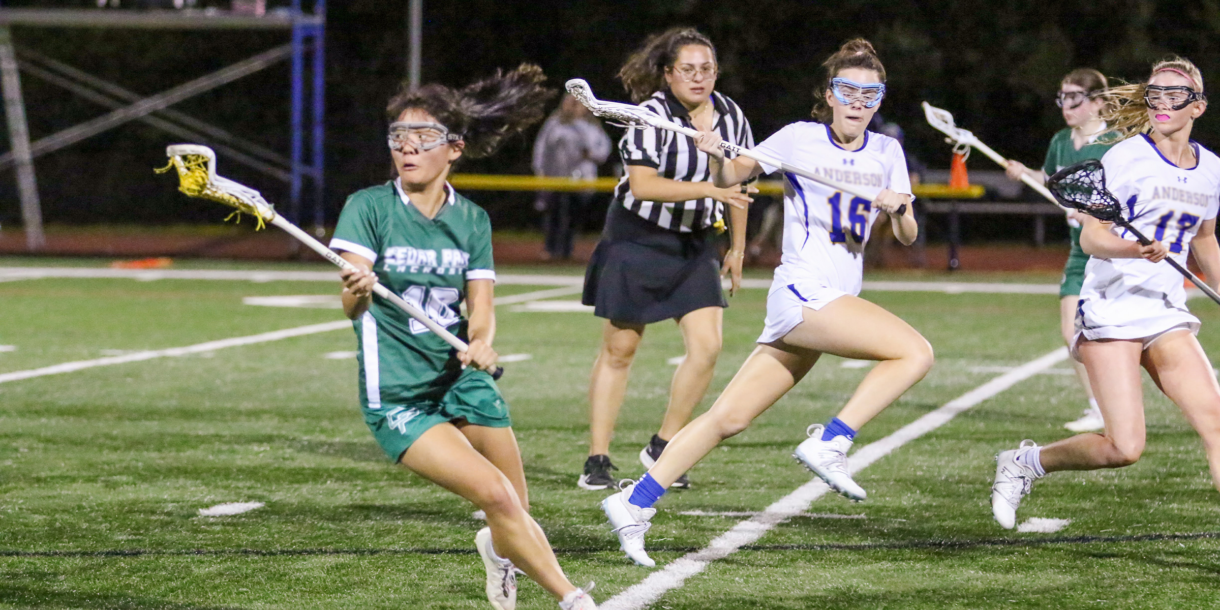 photo of lacrosse officials running while observing the interaction between two lacrosse players; one player has the ball and the other is trying to get in position to check the ball carrier's stick.