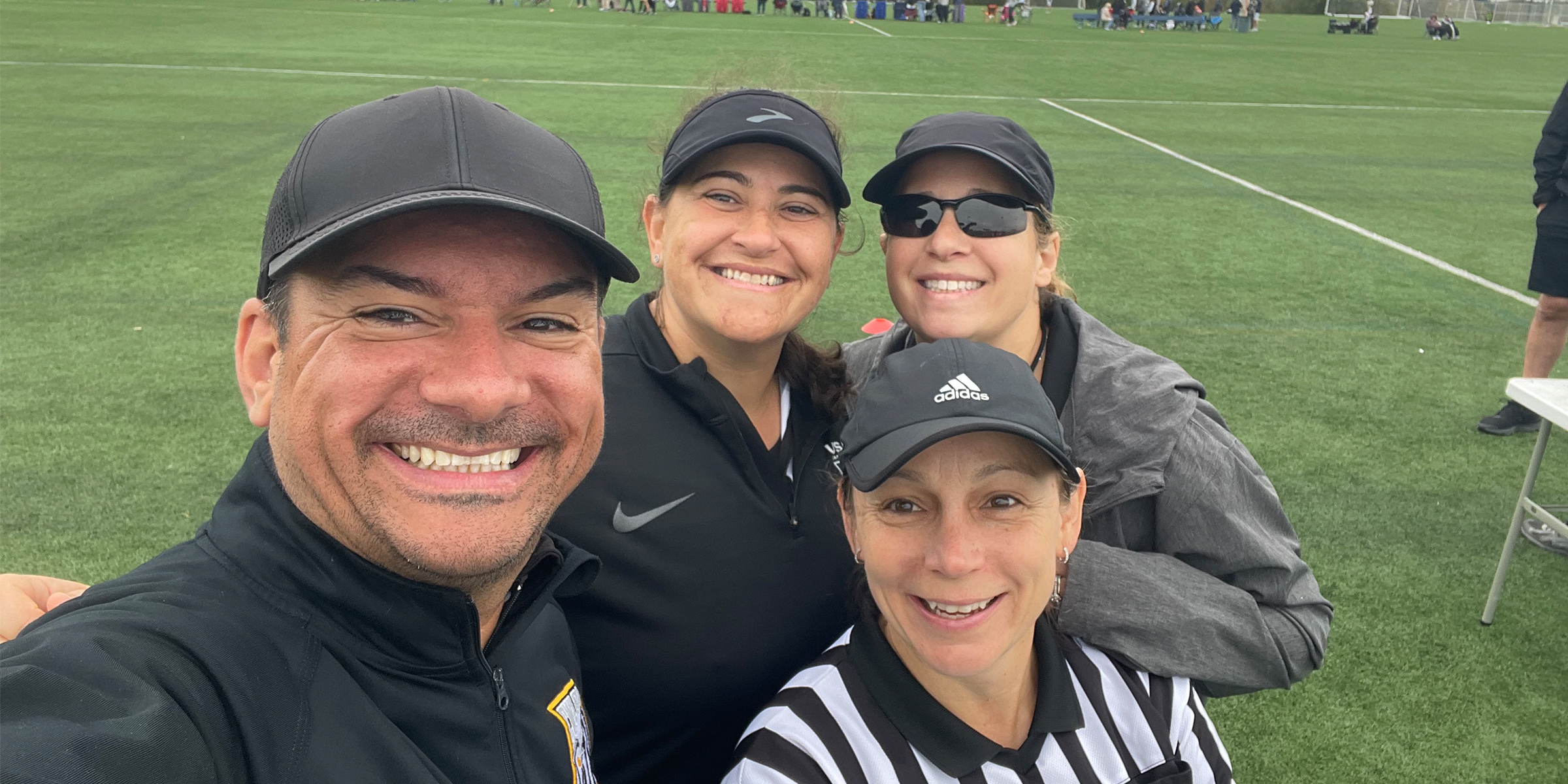 photo of four lacrosse officials in front of a lacrosse field grouped together smiling for the camera